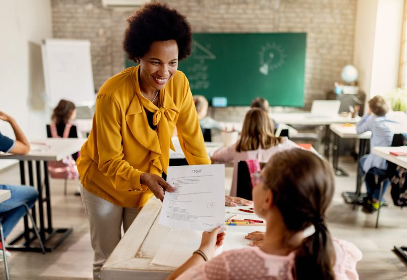 Here are your test results, you got an A! Happy African American teacher giving test results to elementary student in the classroom.